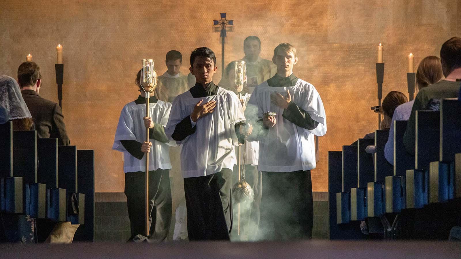 Student carrying a thurible down the center aisle in mass with two students on either side